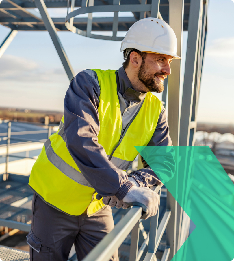 Mann mit Schutzhelm und gelber Warnweste lehnt sich an ein Geländer auf einer Industrieplattform im Freien.
